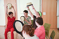 Bei einem Workshop von den Kreativen Bildungstagen. Die Kursleiterin und die Teilnehmenden sitzen auf Sesseln in einem Kreis. Jede und Jeder hat einen Hut in den Händen. Sie strecken die Hüte über sich in die Luft.
