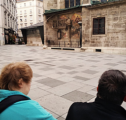 Zwei Personen aus der Gruppe Stehen vor dem Stephansdom und schauen sich die Fasade an. An einer bestimmten Stelle kann man ein Kurzifix sehen. Dahinter sind zwei Wandmalereien. Das Ganze ist von einem kleinen Dachvorsprung vor Regen geschützt.
