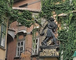 Die Hausfasade beim Griechenbeisl ist mit Efeu überwachsen. An der Wand hängt die Gedenktafel vom lieben Augustin. Darauf sieht man den Augustin mit Dudelsack und fröhlich lachend. Auf der Tafel steht geschrieben: Hier sang sein Lied der Liebe Augustin.