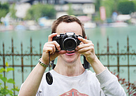 Ein Teilnehmer beim Fotoworkshop. Er steht direkt am Ufer des Wolfgangsees und hält sich die Kamera vors Gesicht um ein Foto zu machen.