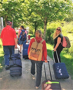 Bei der Ankunft in Strobl am Wolfgangsee. Eine Gruppe von Kursleiterinnen und Teilnehmenden geht mit ihrem Gepäck einen Weg entlang. Um sie herum ist es grün. Die Sonne scheint. Es ist ein sommerlicher Tag. Die Bäume tragen viele grüne Blätter und auch die Wiese am Wegrand ist grün und frisch. 