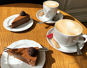Ein hölzener Kaffeehaustisch in gemütlicher Atmosphere. Die Sonne scheint zum Fenster herrein. Auf dem Tisch stehen zwei frische Häferl mit Kaffe und zwei Teller mit jeweils einem leckeren, schokoladigen Stück Sachertorte.