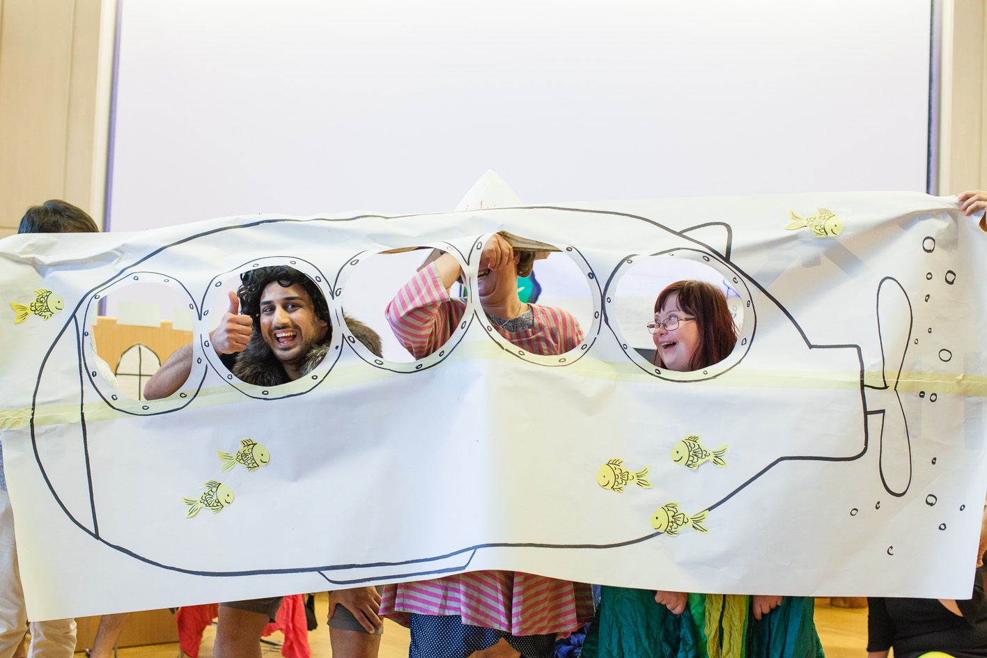 Bei unserem Theaterworkshop haben die Teilnehmerinnen viel Spaß. Drei Frauen halten ein großes Plakat. Auf das Papier wurde ein Uboot gezeichnet. Die Fenster von dem Uboot sind ausgeschnitten. Die Teilnehmerinnen spielen ein Theaterstück. Sie lachen und schauen durch die Luken.