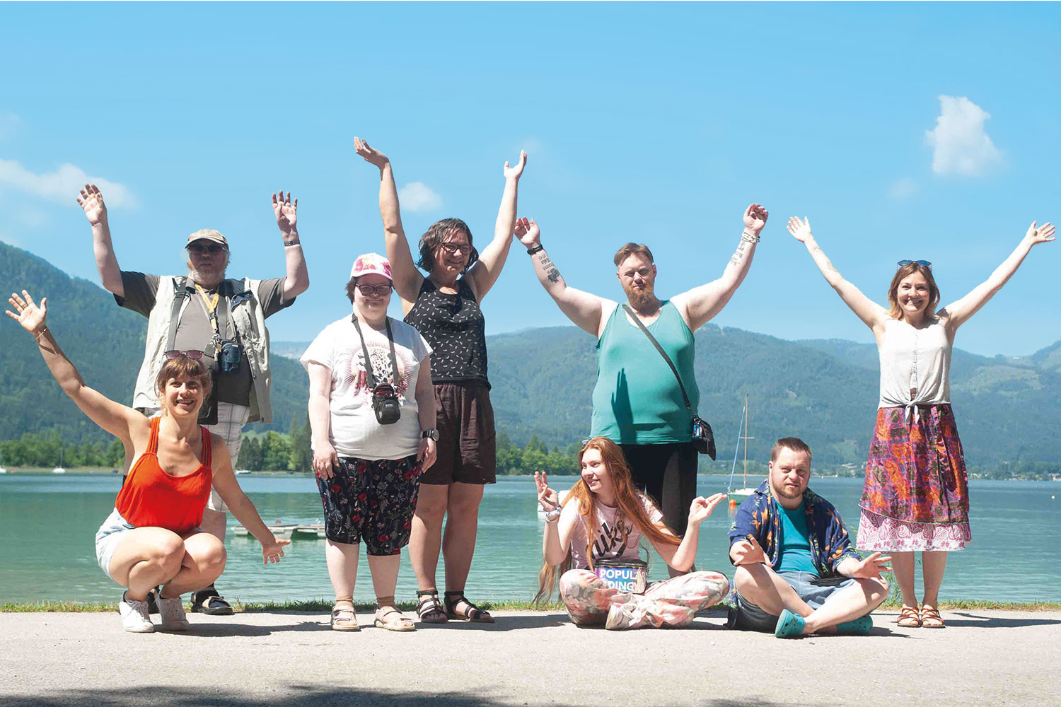 Eine Gruppe 8 Menschen mit und ohne Behinderung stehen am Ufer des Wolfgangsees uns strecken freudig ihre Hände in die Luft. Der Himmel ist blau und die Sonne scheint. Es ist ein wundervoller Frühsommertag.